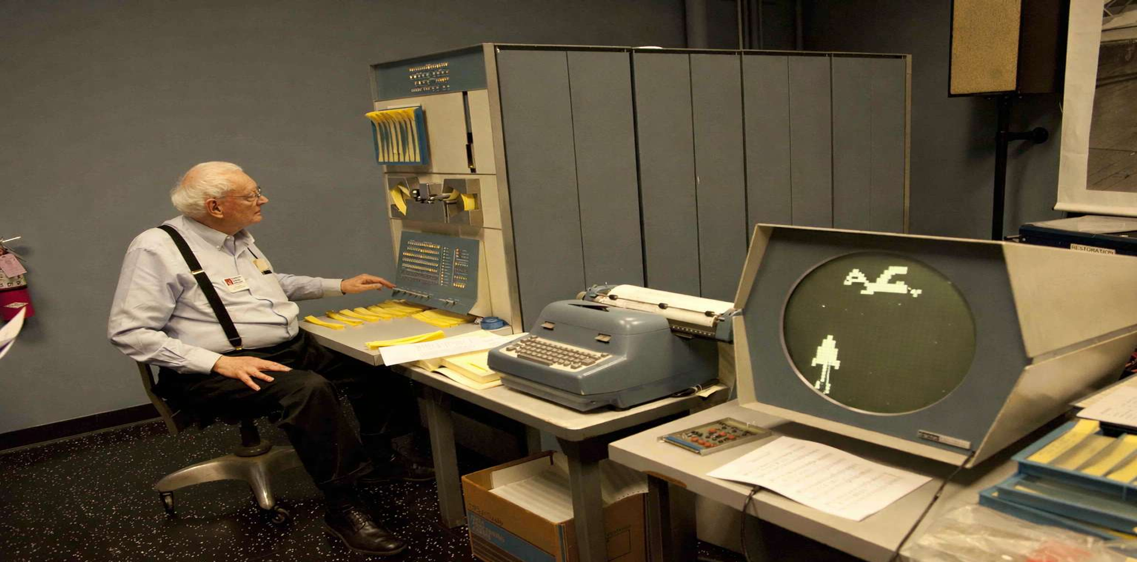 Graphic of an elderly man seated at a vintage mainframe computer, operating a console with punch cards, a typewriter terminal, and a CRT screen displaying simple graphics in a computer lab or museum setting featured in hyperfast herminator
